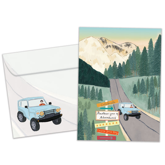 Front view of a Birthday greeting card featuring an illustration of a car driving on a road through the mountains, with a matching envelope.
