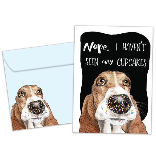 Front view of a birthday greeting card featuring an illustration of a dog’s face denying eating the cupcakes, with a matching envelope.
