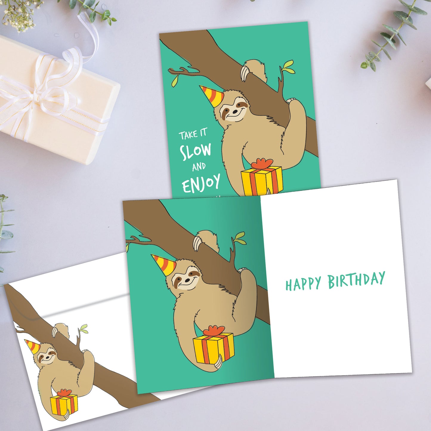 Front and inside view of a birthday card showcasing a sloth holding a present and the message "Take it slow", with a matching envelope.