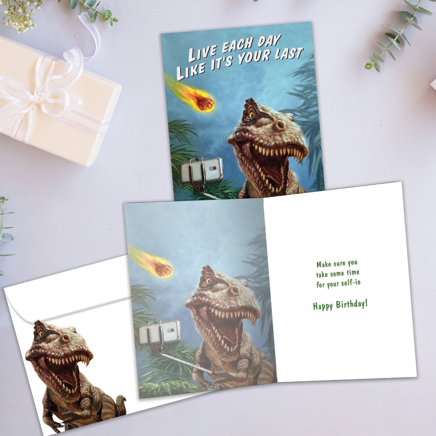 Front and inside view of a Birthday greeting card showcasing a dinosaur taking a selfie with a fireball flying towards it, with a matching envelope.