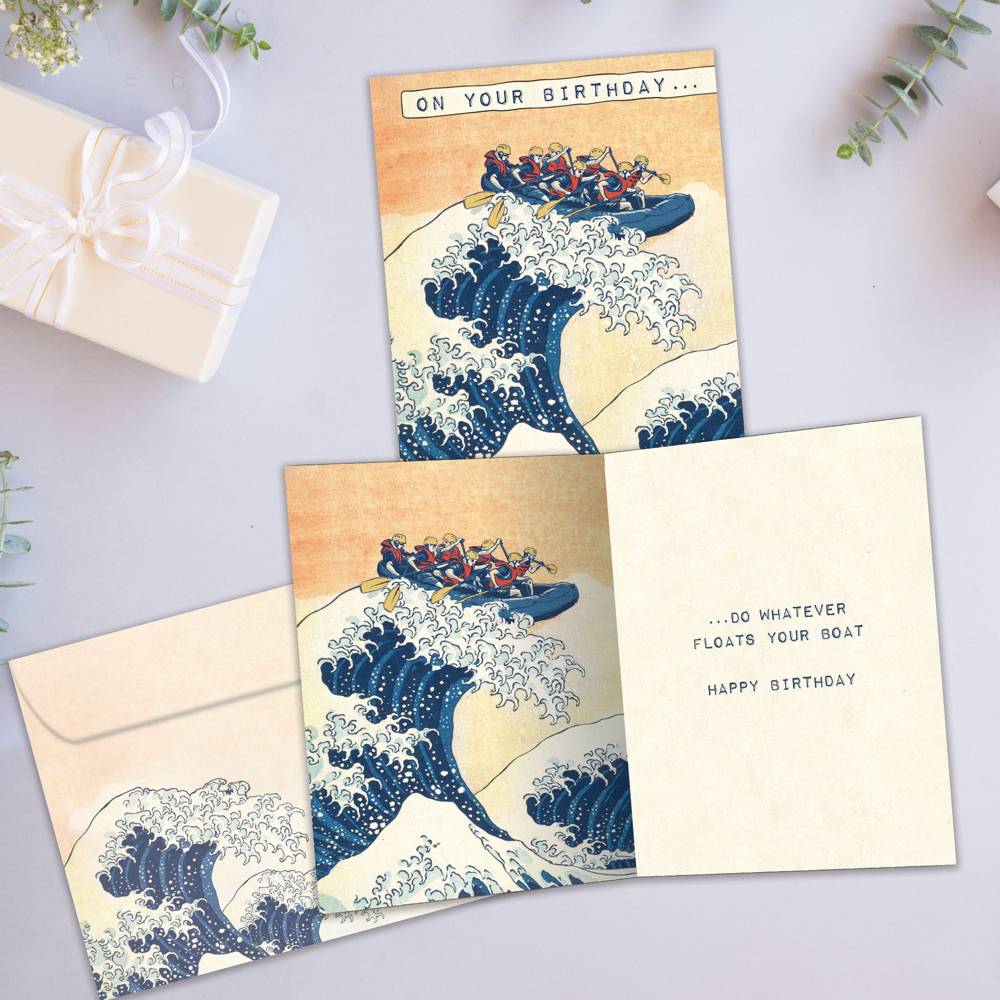 Front and inside view of a Birthday greeting card showcasing an illustration of friends in a raft paddling on a giant wave, with a matching envelope.