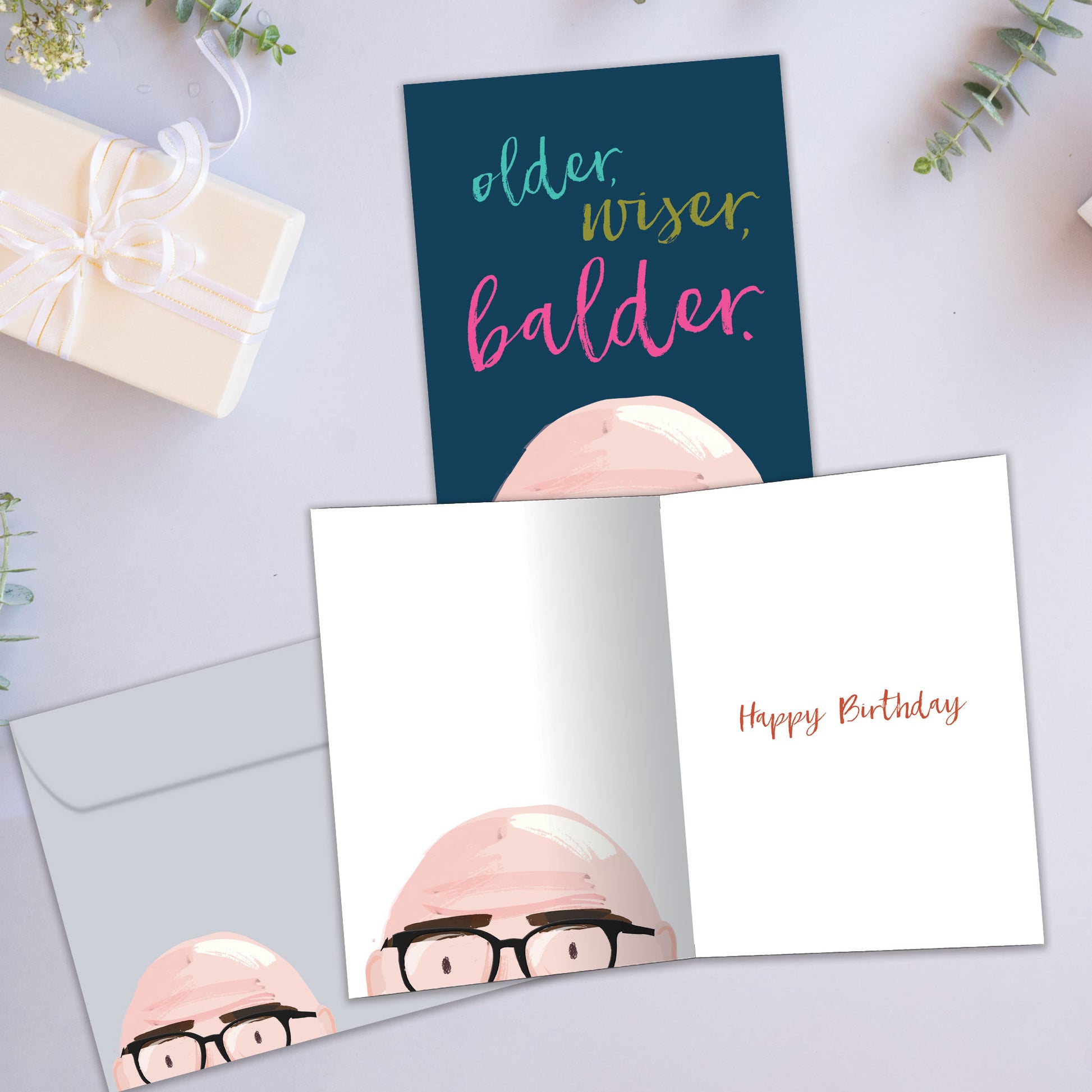 Front and inside view of a Birthday greeting card showcasing the top of a bald man's head with the words "older, wiser, balder", with a matching envelope.