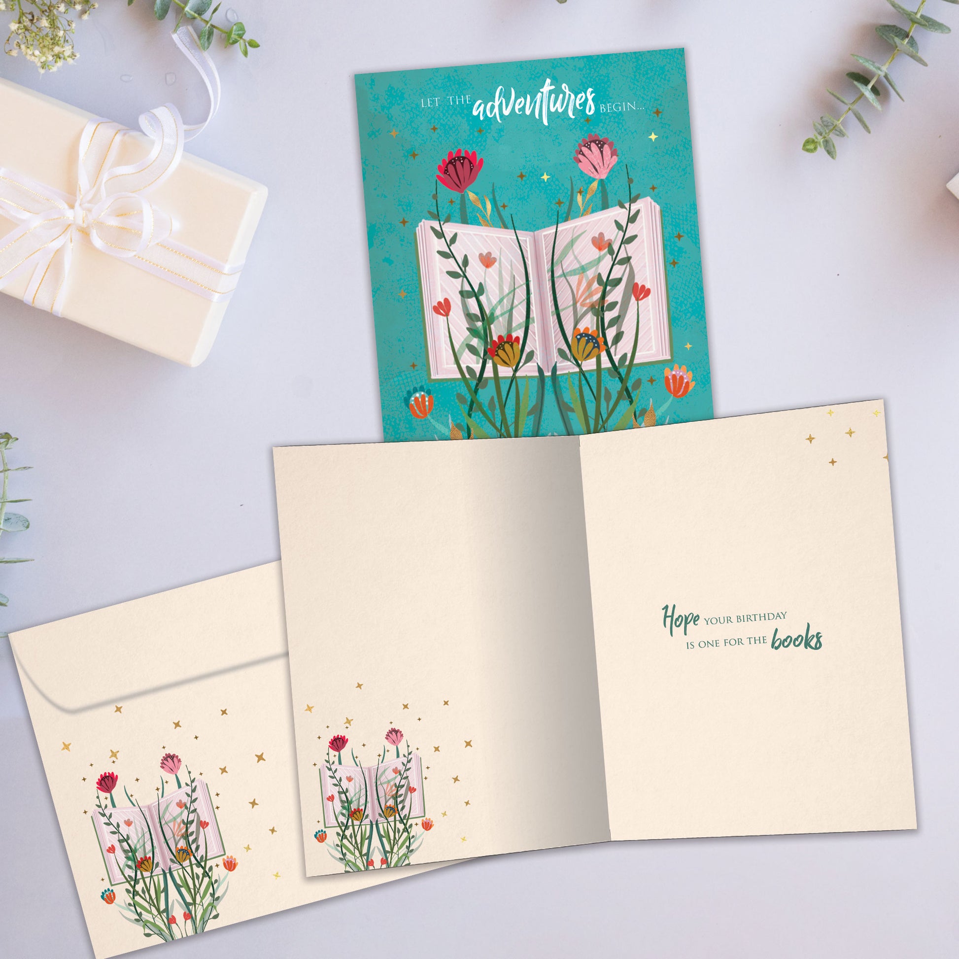 Front and inside view of a Birthday greeting card showcasing an open book with flowers and a message about adventures, with a matching envelope.