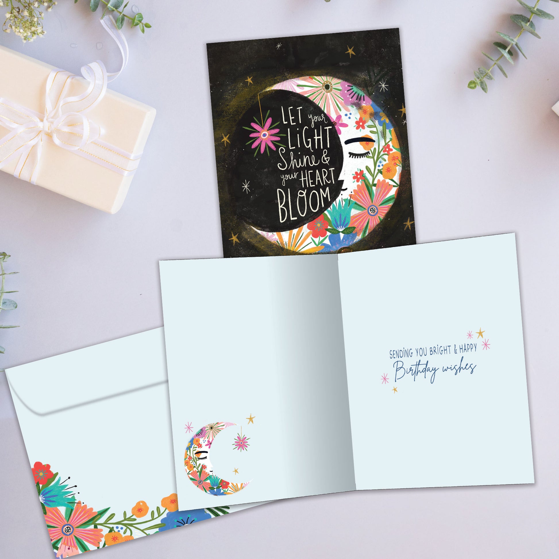 Front and inside view of a birthday greeting card showcasing an illustration of a floral crescent moon with a message about letting your light shine, with a matching envelope.