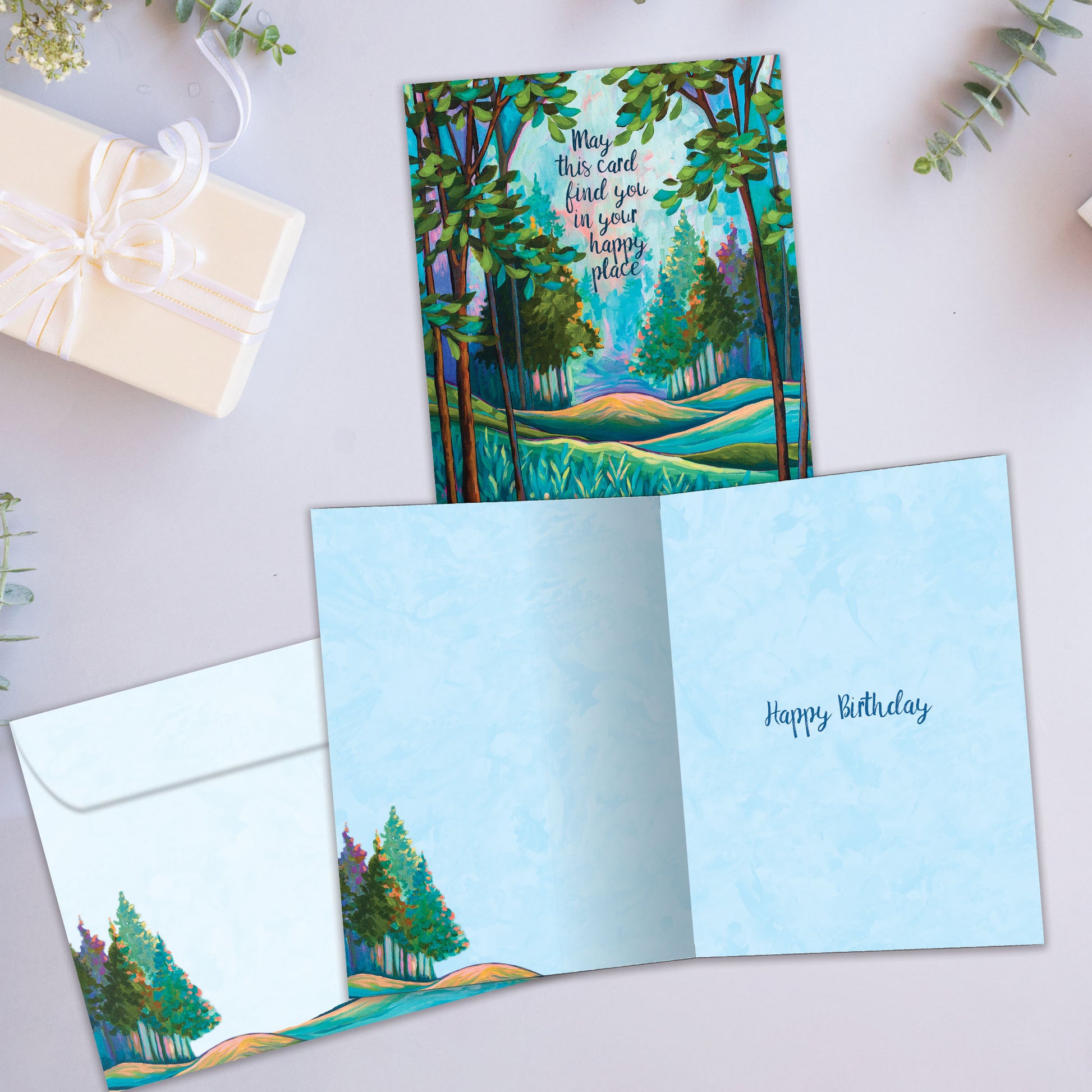 Front and inside view of a birthday greeting card showcasing an illustration of a tranquil forest with a message about finding your happy place, with a matching envelope.