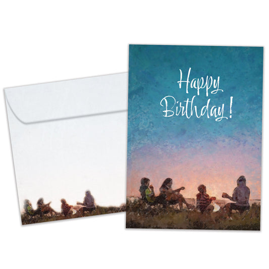 Front view of a birthday greeting card featuring an illustration of a family sitting and playing guitar around a campfire with the message "Happy Birthday!", with a matching envelope.