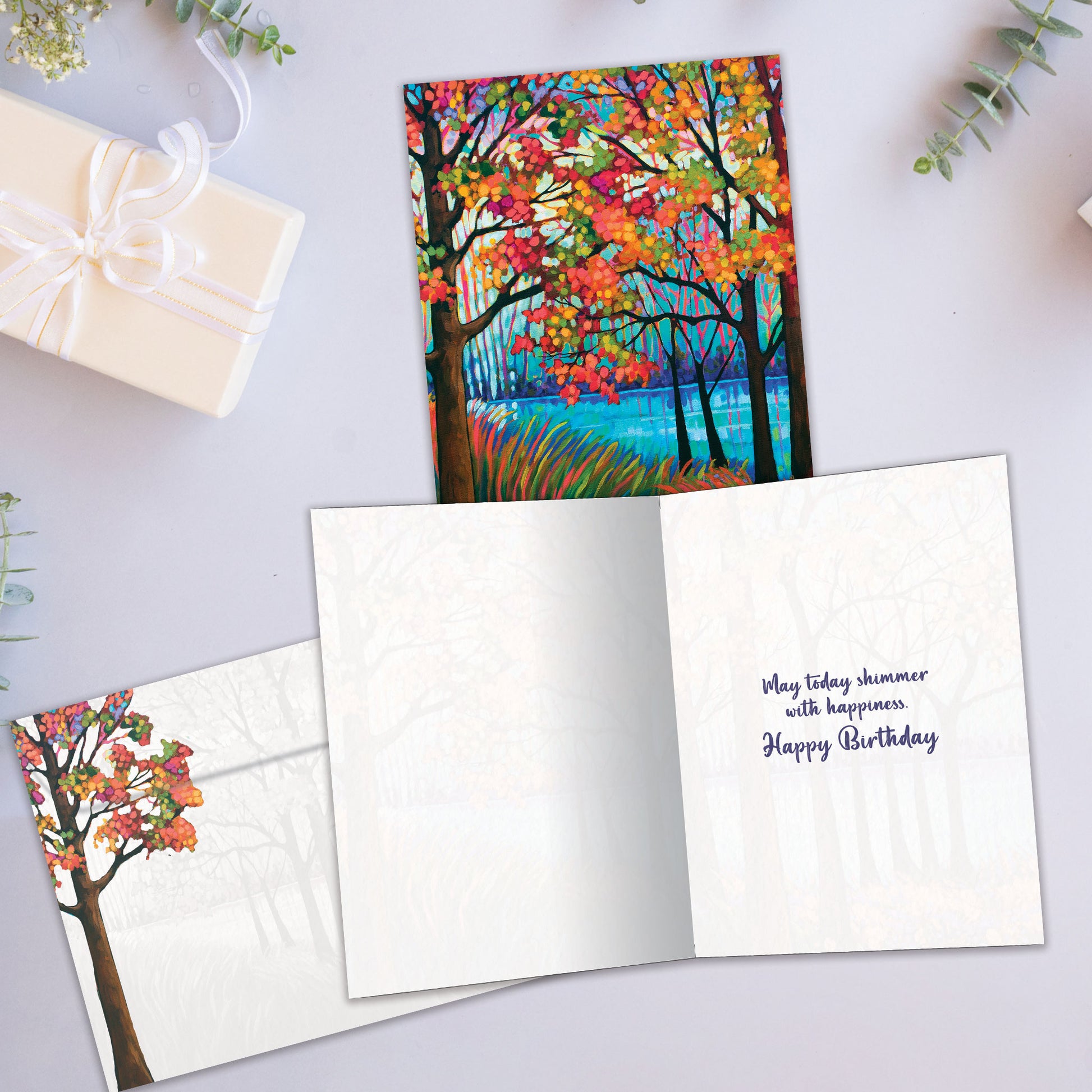 Front and inside view of a birthday greeting card showcasing a vibrant illustration of fall trees and a shimmering lake, with a matching envelope.