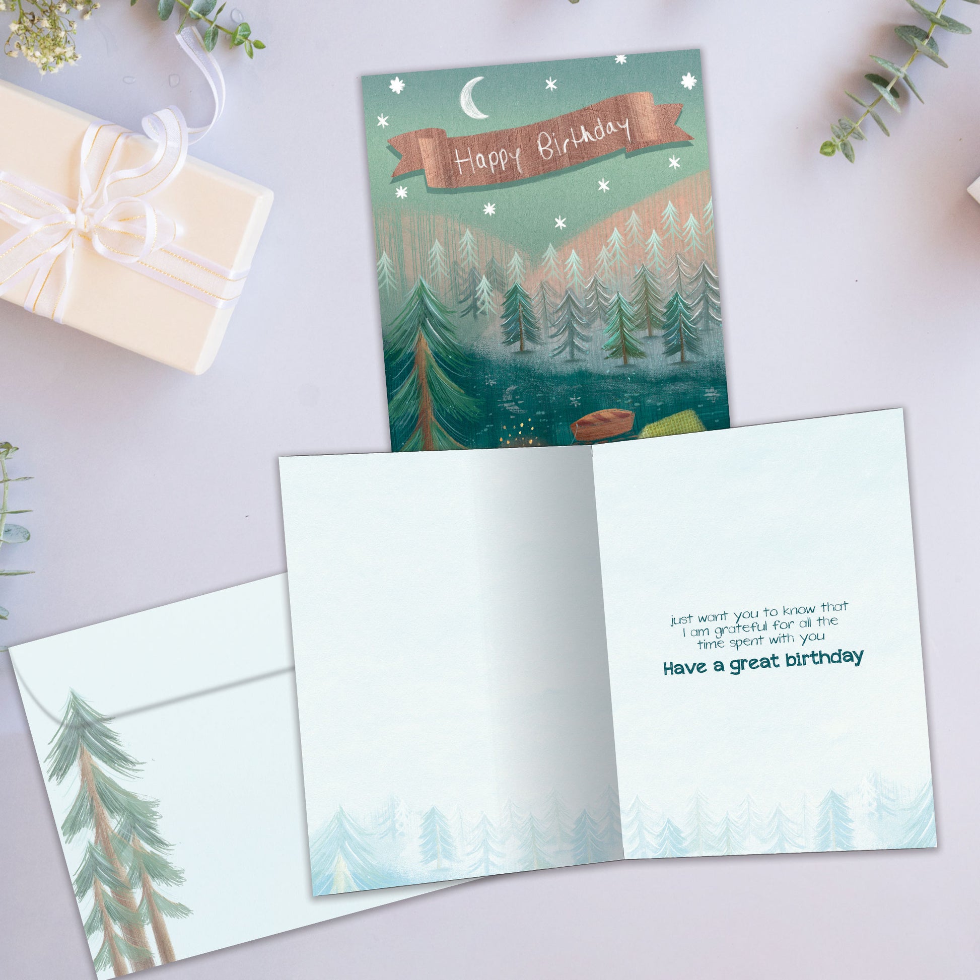Front and inside view of a birthday greeting card showcasing an illustration of two people sitting by a campfire, a tent, and a forest with the message "Happy Birthday", with a matching envelope.