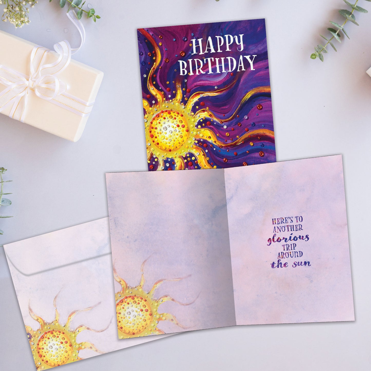 Front and inside view of a birthday greeting card showcasing an abstract illustration of a sun on a purple background with the words "Happy Birthday", with a matching envelope.