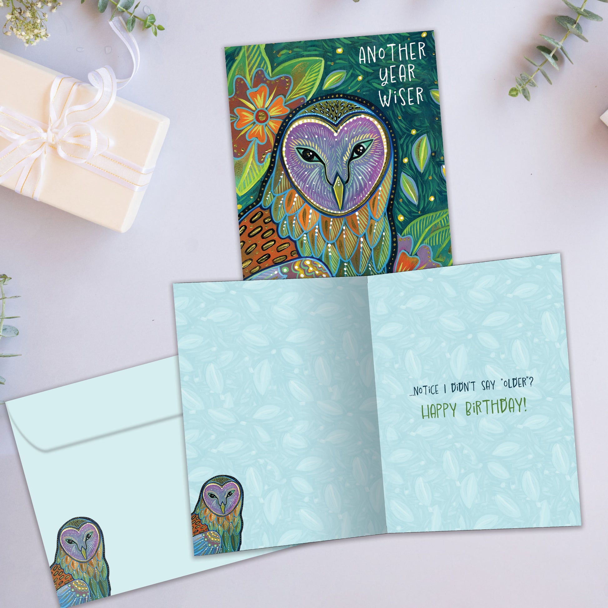 Front and inside view of a birthday greeting card showcasing an artistic and colourful illustration of an owl and foliage with the message "another year wiser", with a matching envelope.