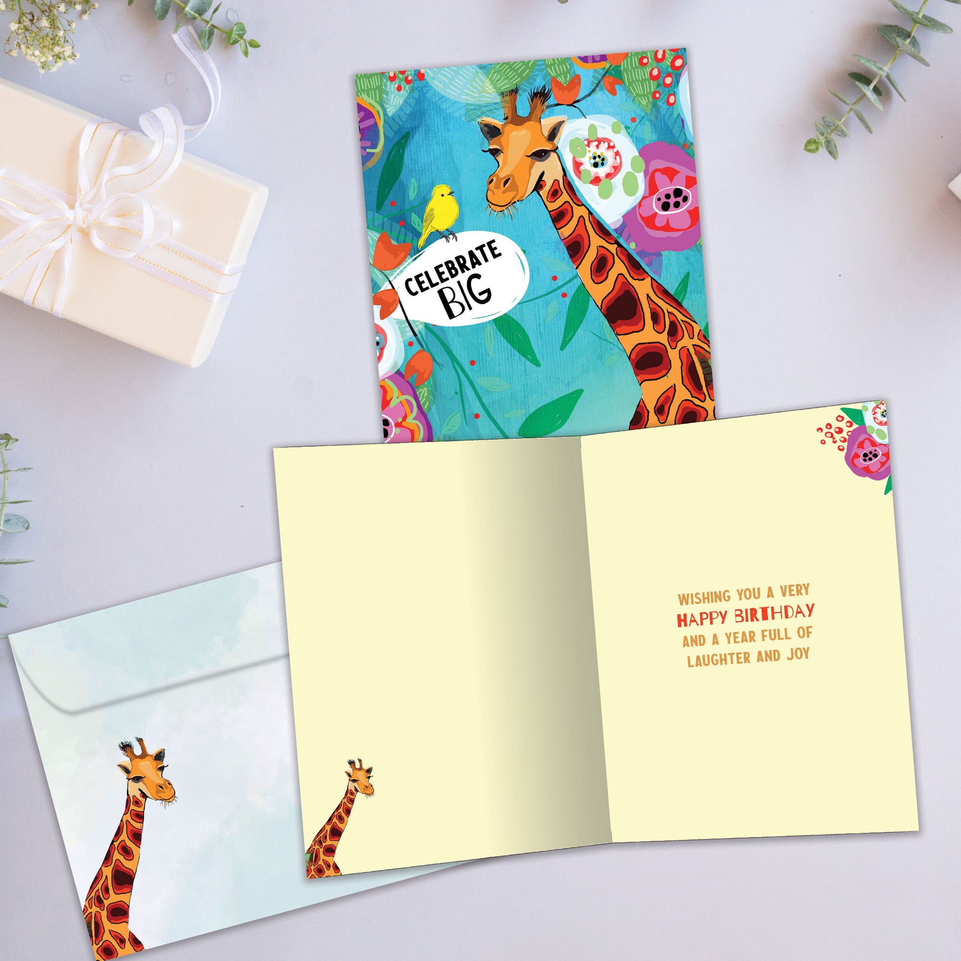 Front and inside view of a birthday greeting card showcasing a colourful illustration of a giraffe and bird in primary colours with the words "celebrate big", with a matching envelope.