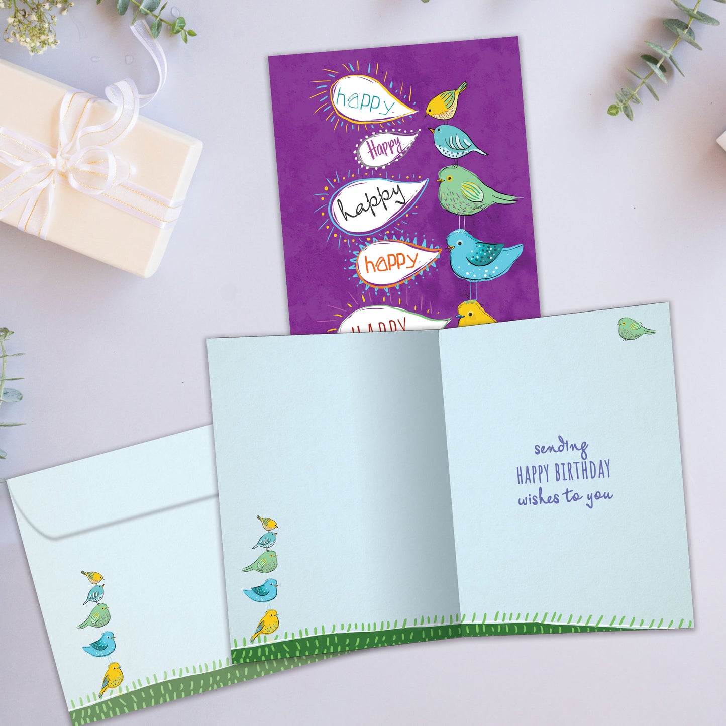 Front and inside view of a birthday greeting card showcasing an illustration of colourful birds standing on top of each other with speech bubbles saying "happy", with a matching envelope.