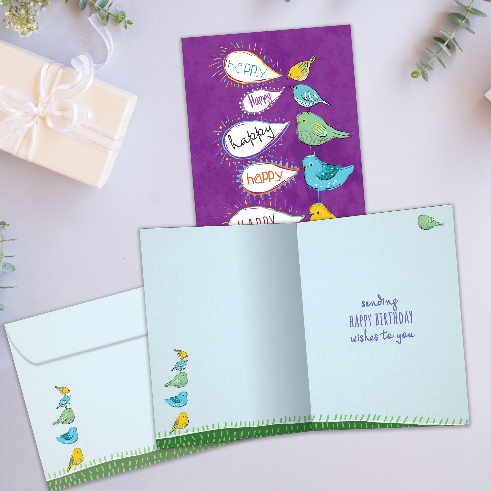 Front and inside view of a birthday greeting card showcasing an illustration of colourful birds standing on top of each other with speech bubbles saying "happy", with a matching envelope.