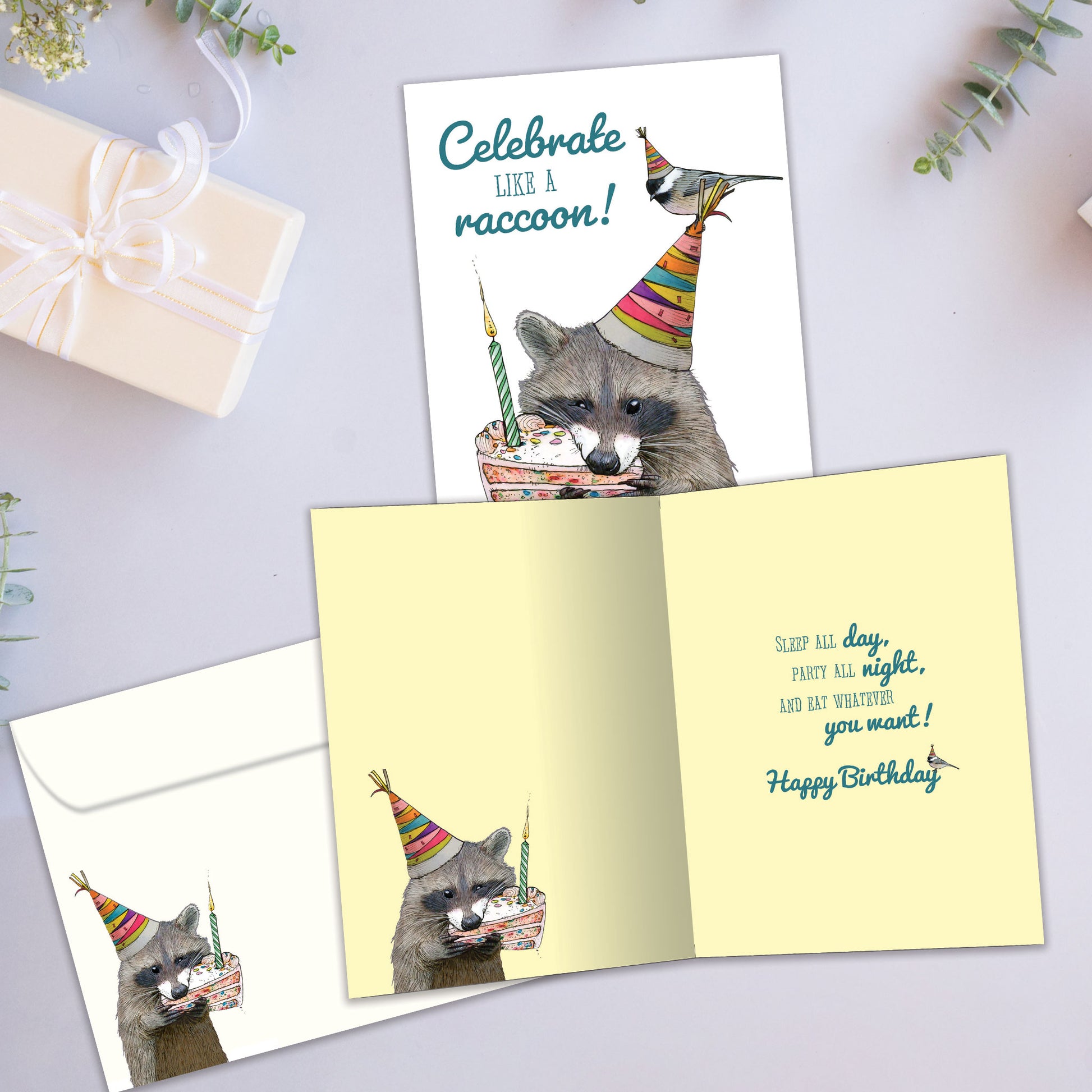 Front and inside view of a Birthday greeting card showcasing a raccoon eating cake with the message "Celebrate like a raccoon!", with a matching envelope.