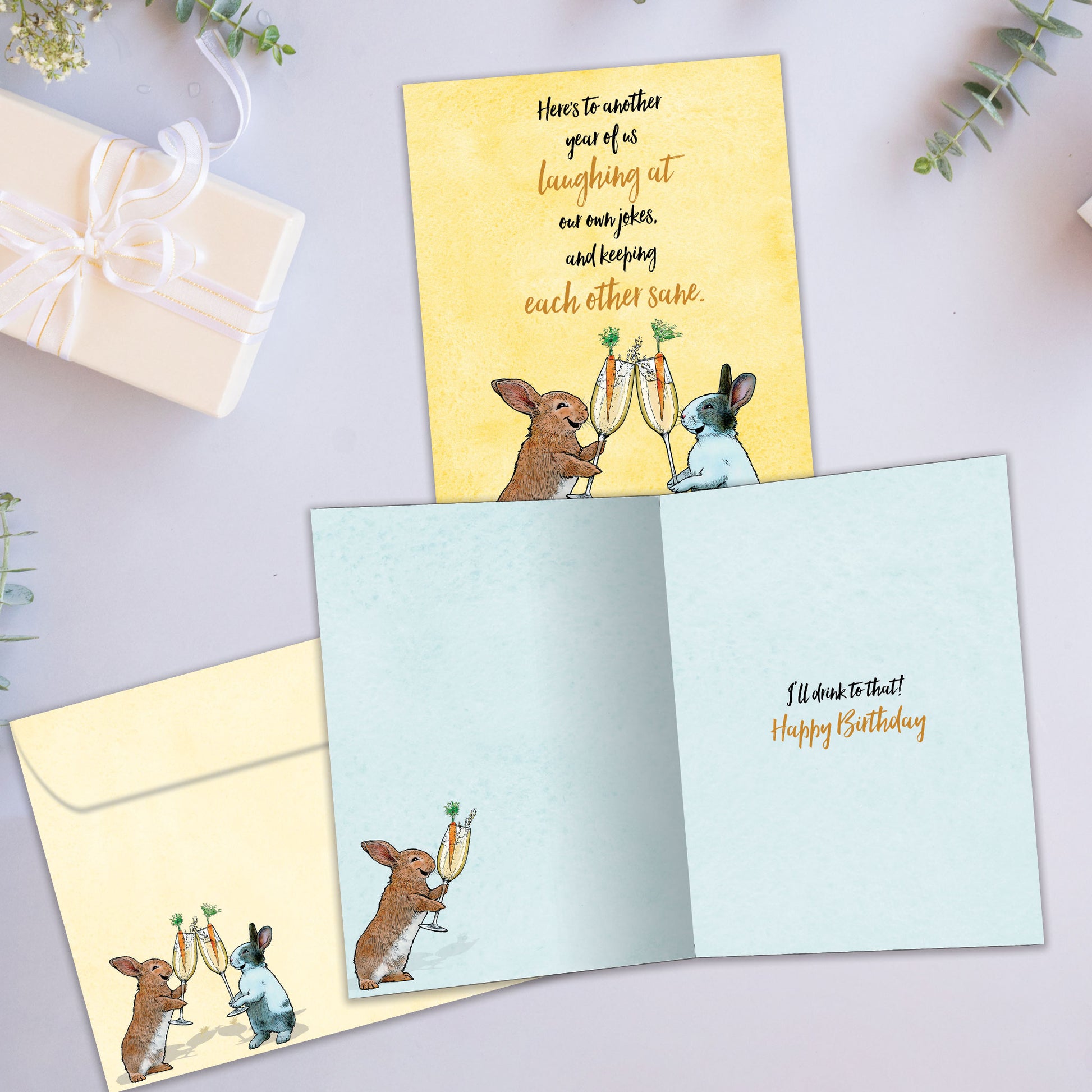 Front and inside view of a Birthday greeting card showcasing two rabbits with champagne glasses and a message about “us”, with a matching envelope.