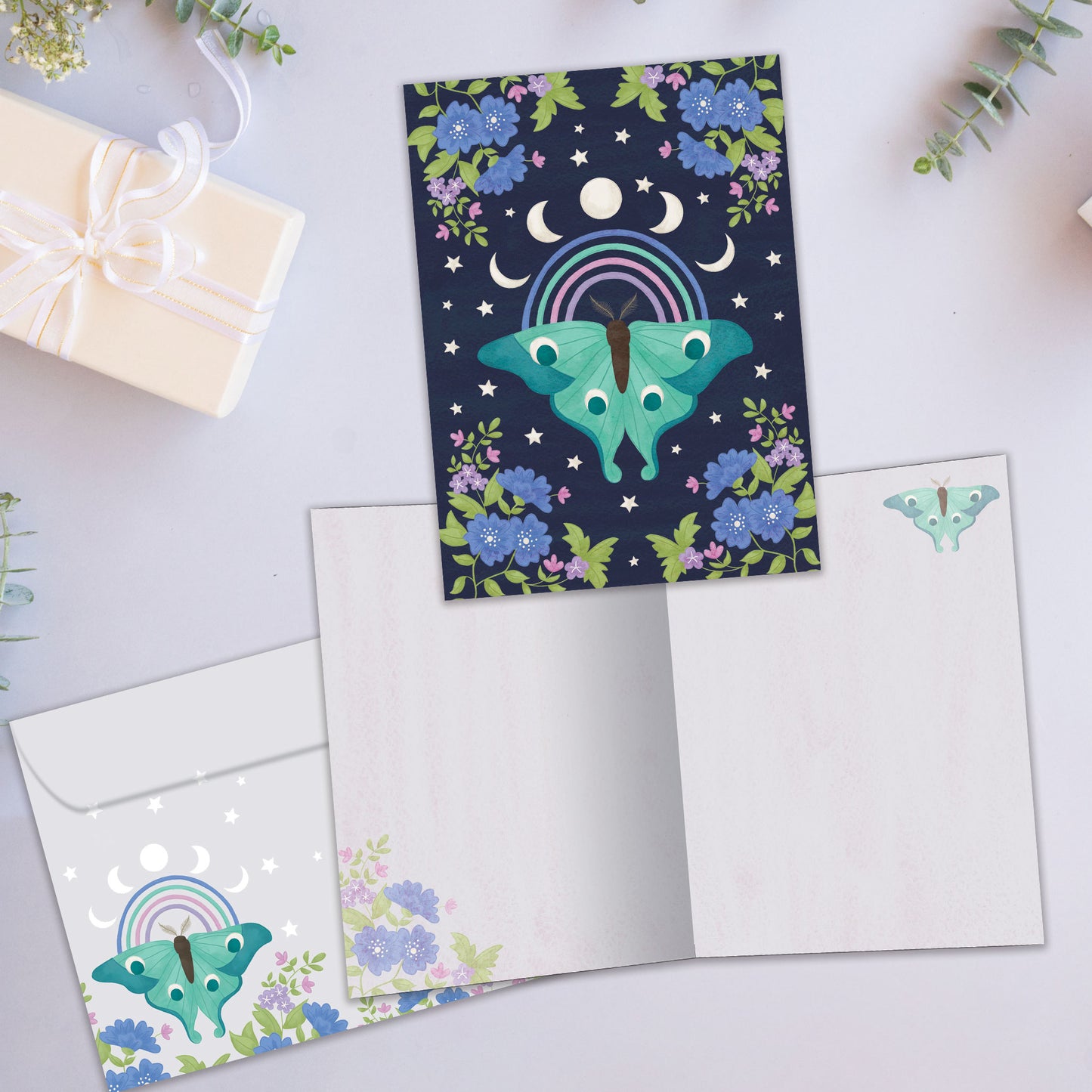 Front and inside view of an All Occasion greeting card showcasing a detailed moth illustration with moon phases and flowers on a dark blue background, with a matching envelope.