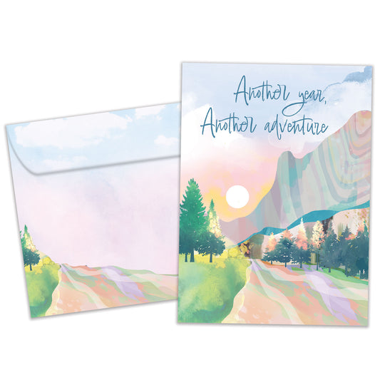 Front view of a Birthday greeting card featuring an illustration of a country road with the message "Another year, Another adventure," with a matching envelope.