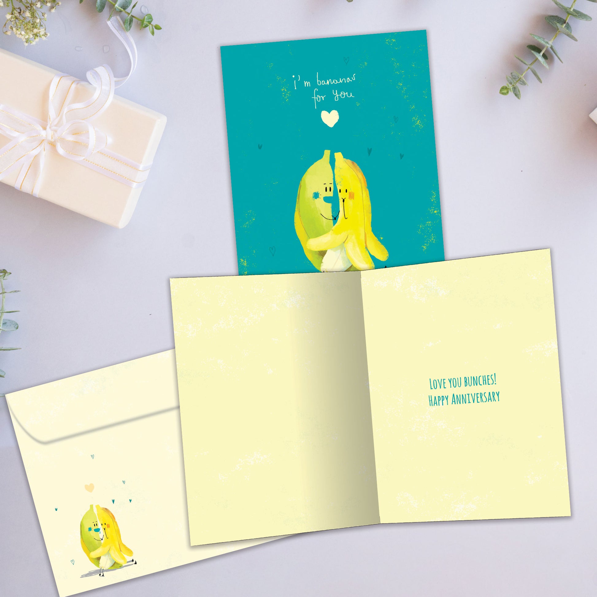 Front and inside view of an Anniversary greeting card showcasing two bananas hugging on a teal background with a loving message, with a matching envelope.