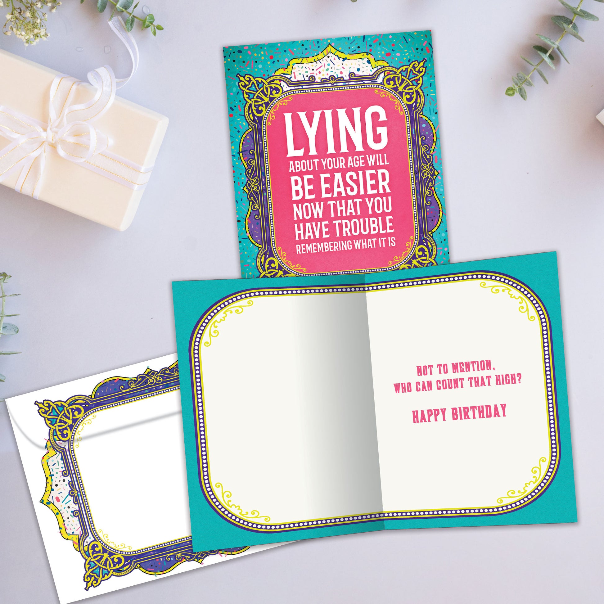 Front and inside view of a Birthday greeting card showcasing a funny message about lying about your age on a pink and teal background, with a matching envelope.